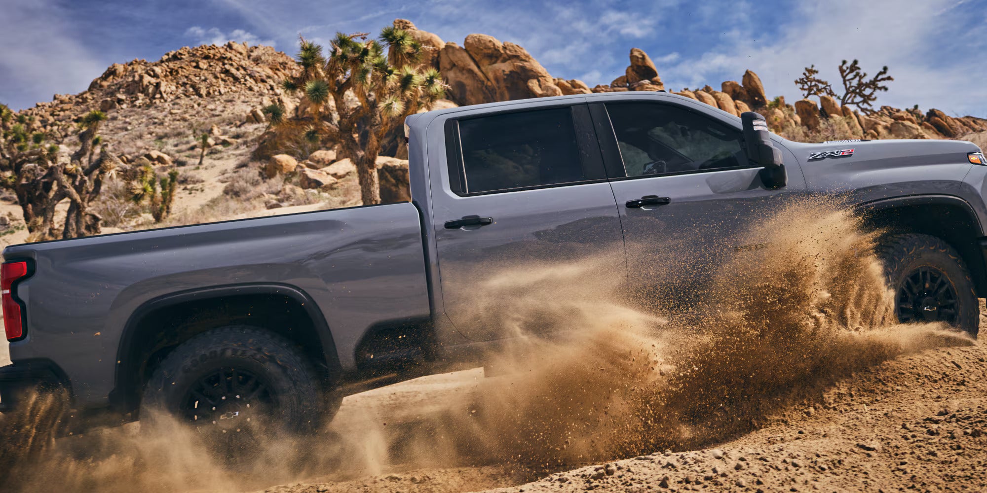 a gray 2025 silverado hd driving in sand with a rocky desert background