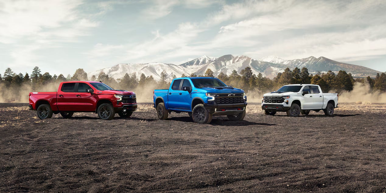 Three 2025 Chevrolet Silverado trucks lined up in a scenic outdoor setting with mountains, displaying model variety.
